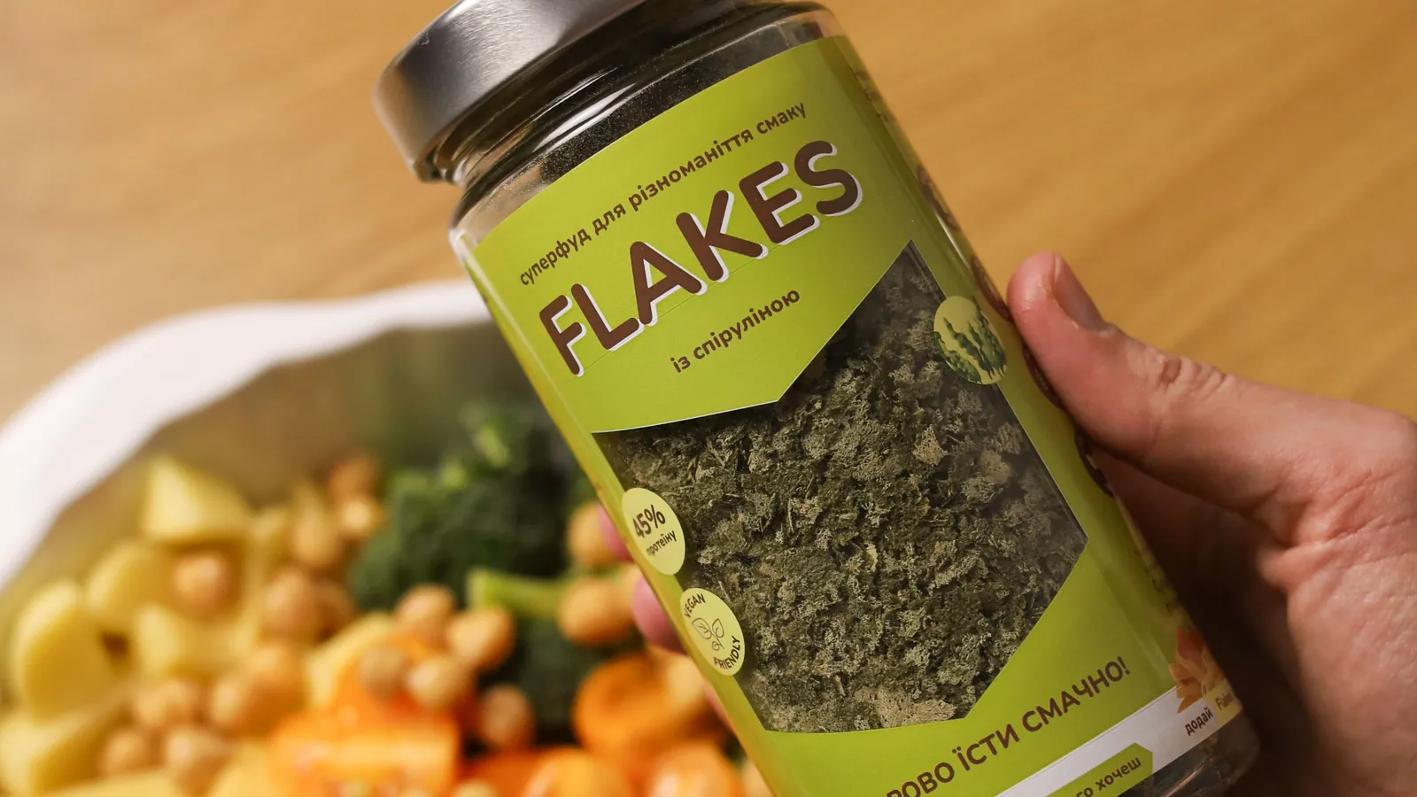 Рука тримає банку Flakes зі спіруліною над мискою з картоплею, нутом і броколі